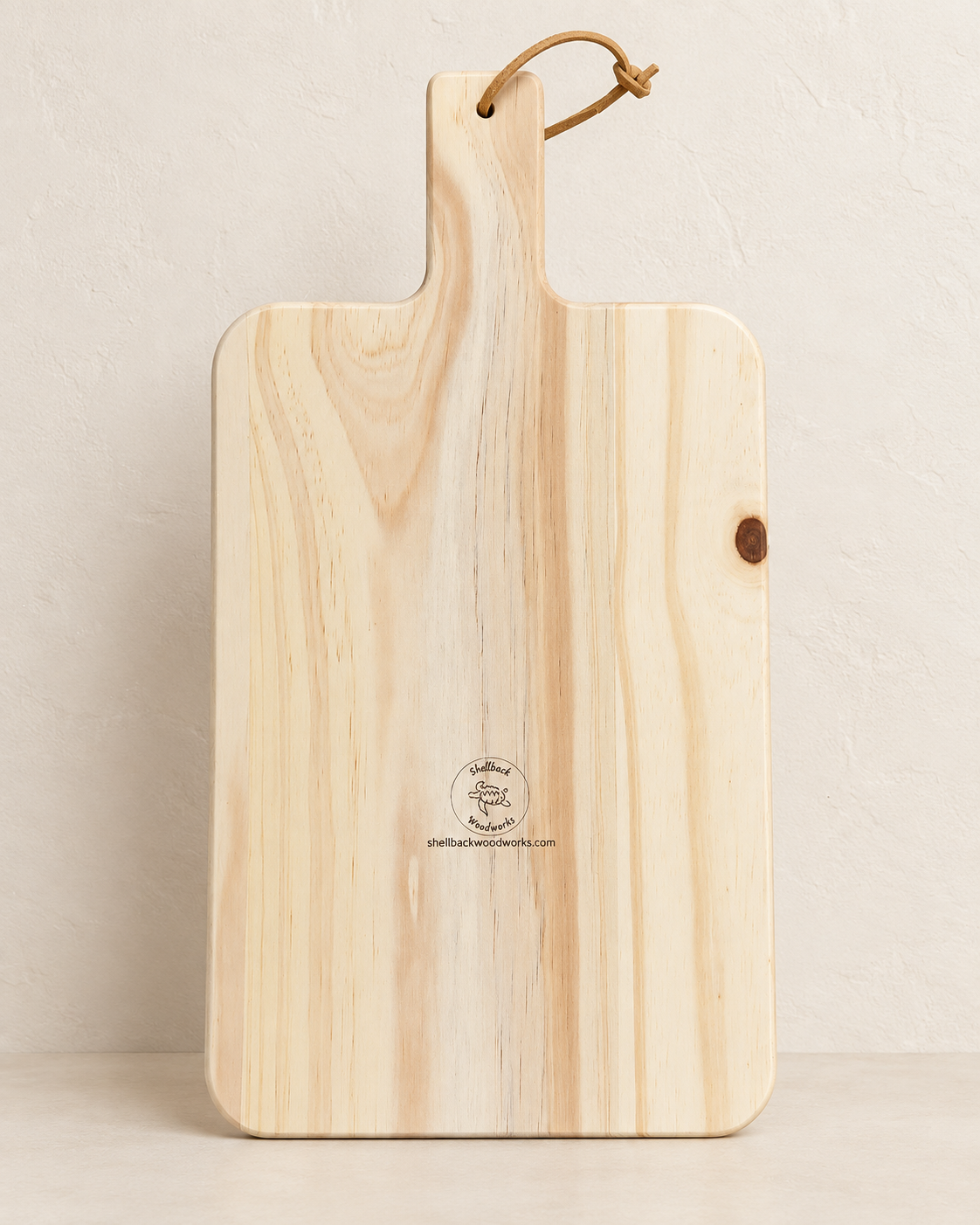 Back of natural pine kitchen paddle board with leather tie, Shellback Woodworks mark, and website